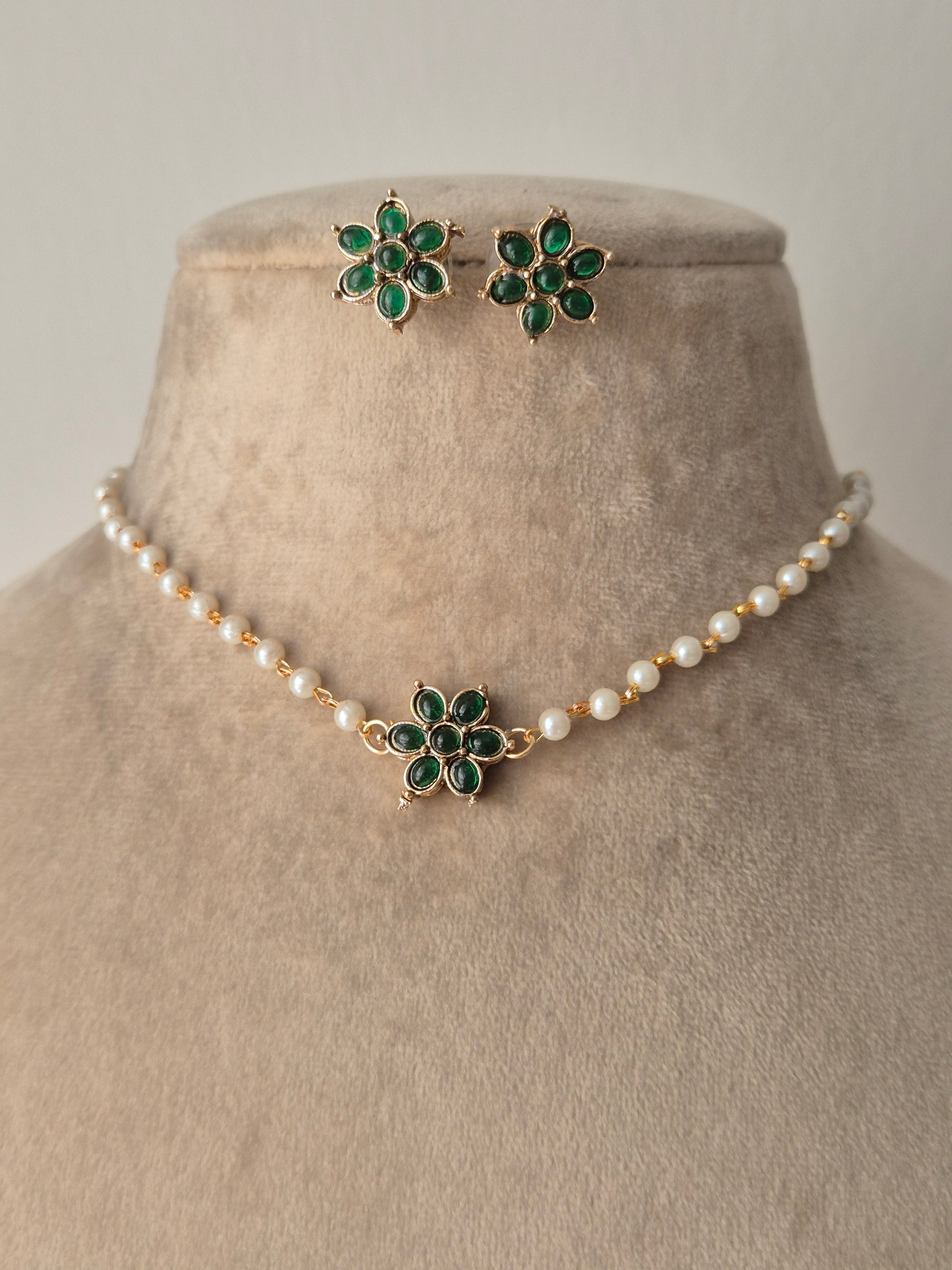 A delicate pearl strand choker featuring a central gold-plated floral motif with green stones, shown with matching small floral stud earrings by naitri.in