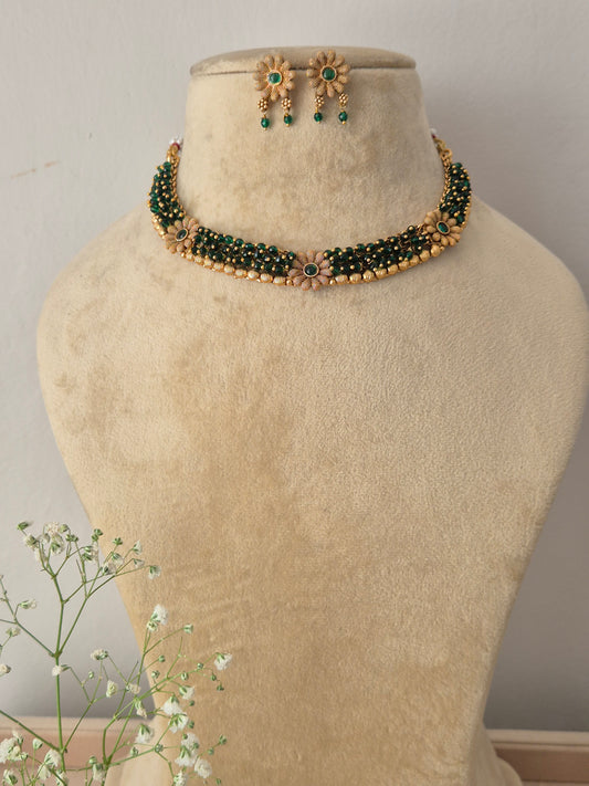A gold-plated multi-strand choker necklace featuring dark green beads and a central gold-trimmed floral motif, displayed on a beige mannequin bust with matching hanging earring by naitri.in