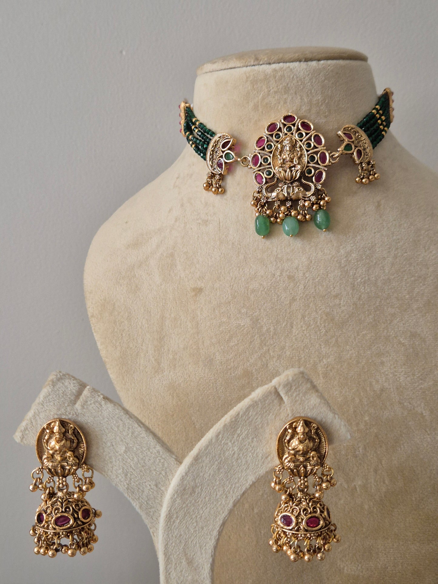 Traditional Indian bridal choker set in gold finish with emerald green beads and ruby pink stones, ideal for festive and wedding wear Antique gold choker necklace set with green beads and Goddess Lakshmi motif pendant, including matching drop earrings by naitri.in