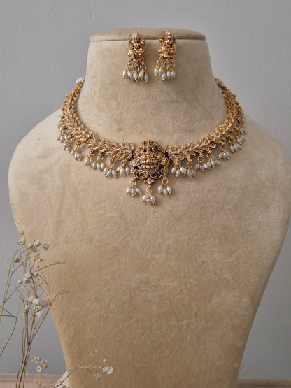 An antique gold-plated temple jewelry set featuring a Goddess Lakshmi central motif, intricate floral carvings, and delicate white pearl droplets, including matching Jhumka-style earrings by naitri.in