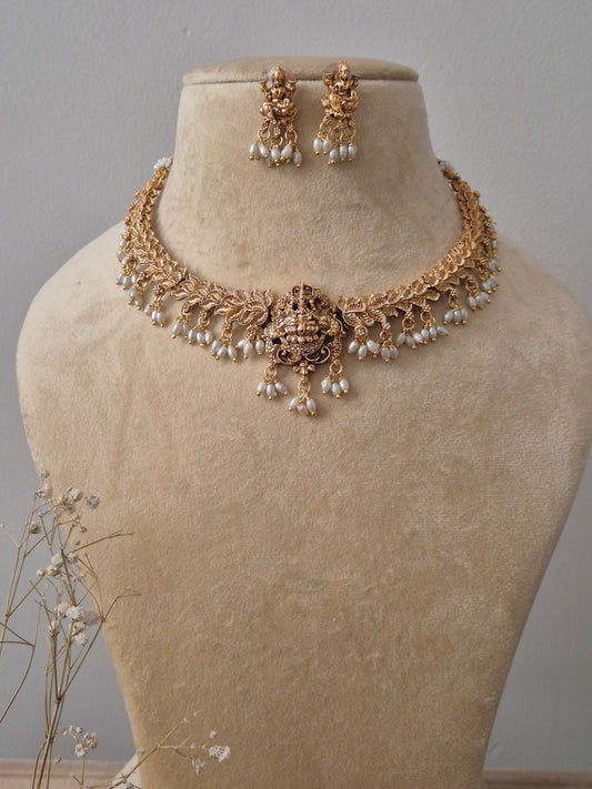 An antique gold-plated temple jewelry set featuring a Goddess Lakshmi central motif, intricate floral carvings, and delicate white pearl droplets, including matching Jhumka-style earrings by naitri.in