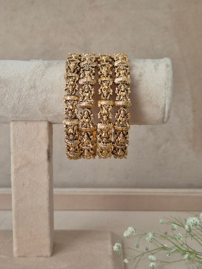 A wide, chunky gold cuff bracelet with intricate openwork floral and scroll patterns, Four traditional gold bangles with detailed filigree work and god/goddess relief carving on a neutral-toned by naitri.in