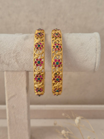 Set of 2 gold-plated bangles featuring delicate floral jaal scrollwork studded with ruby red and emerald green stones by naitri.in