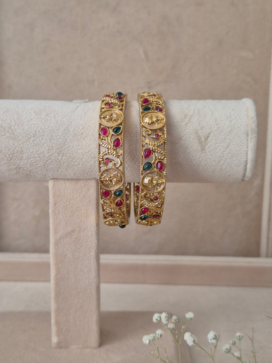 Set of 2 Chirasvi gold bangles by Naitri featuring intricate traditional engravings, accented with alternating ruby-red and emerald-green gemstones, by naitri.in