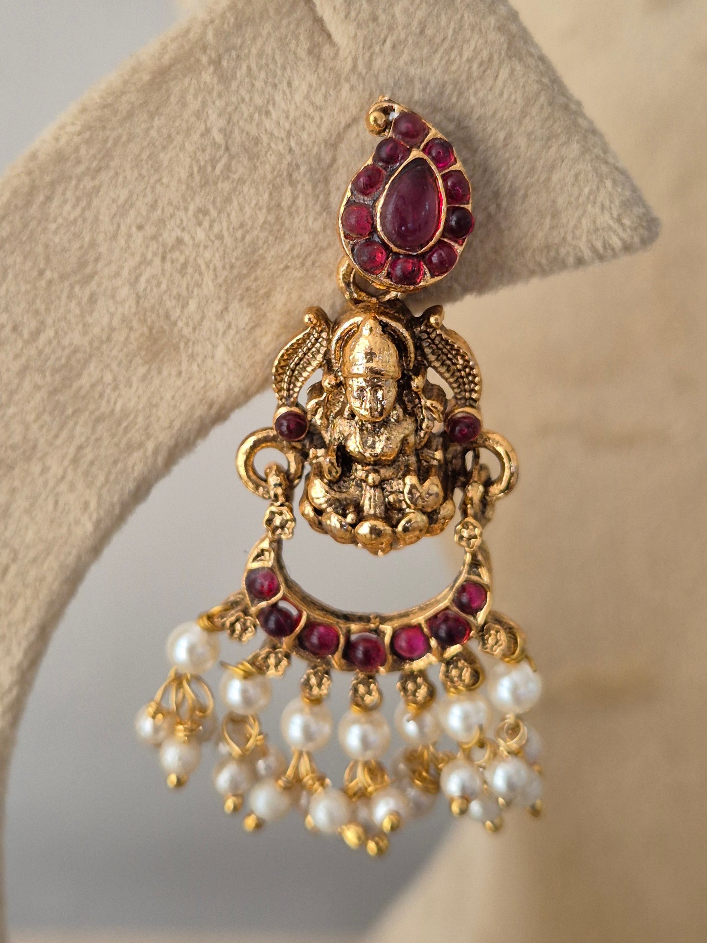 An ornate gold-toned earring piece featuring a central deity motif, a teardrop-shaped purple gemstone, and a fan of white faux pearls accented with small gold beads, by naitri.in