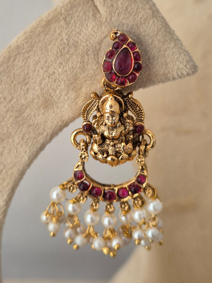 An ornate gold-toned earring piece featuring a central deity motif, a teardrop-shaped purple gemstone, and a fan of white faux pearls accented with small gold beads, by naitri.in
