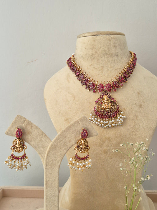 A traditional Indian jewelry set, featuring a choker necklace and matching dangle earrings with gold-plated motifs, deep pink stones, and white faux pearls by naitri.in