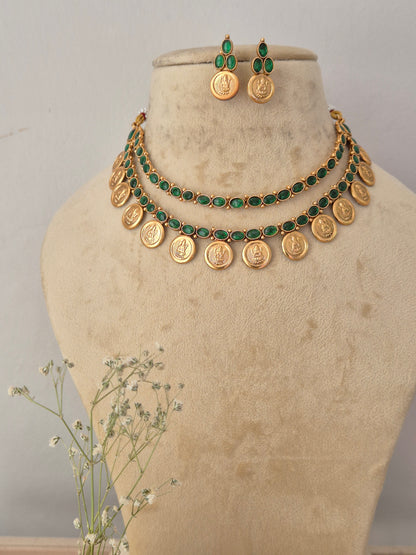 Gold coin necklace and matching drop earrings set displayed on a beige jewelry bust. The necklace features a row of vibrant green emerald-style stones leading to a fringe of hanging circular gold coins, with matching green stone and gold coin earrings by naitri.in