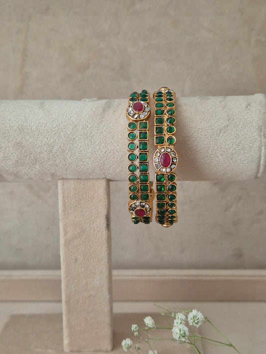 A pair of gold-plated bangle bracelets decorated with green square-cut stones and oval pinkish-red center stones, by naitri.in