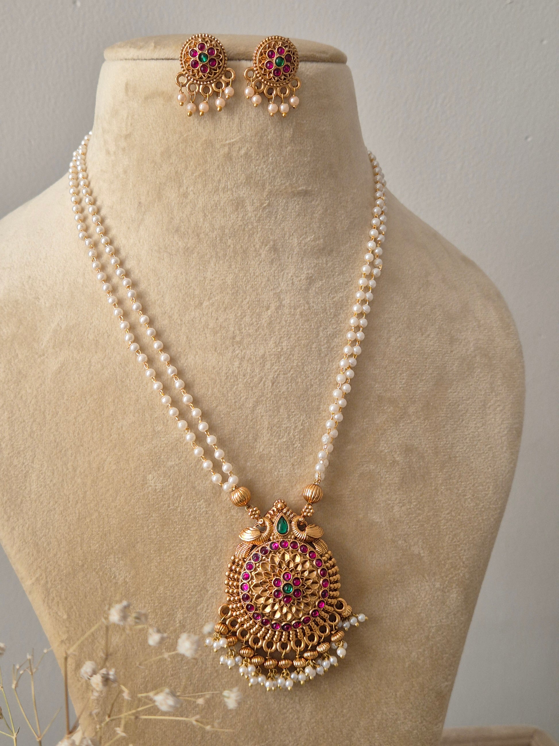 A two-row white pearl strand necklace with a central gold-toned medallion pendant featuring intricate floral carvings, pink gemstones, and white bead droplets by naitri .in