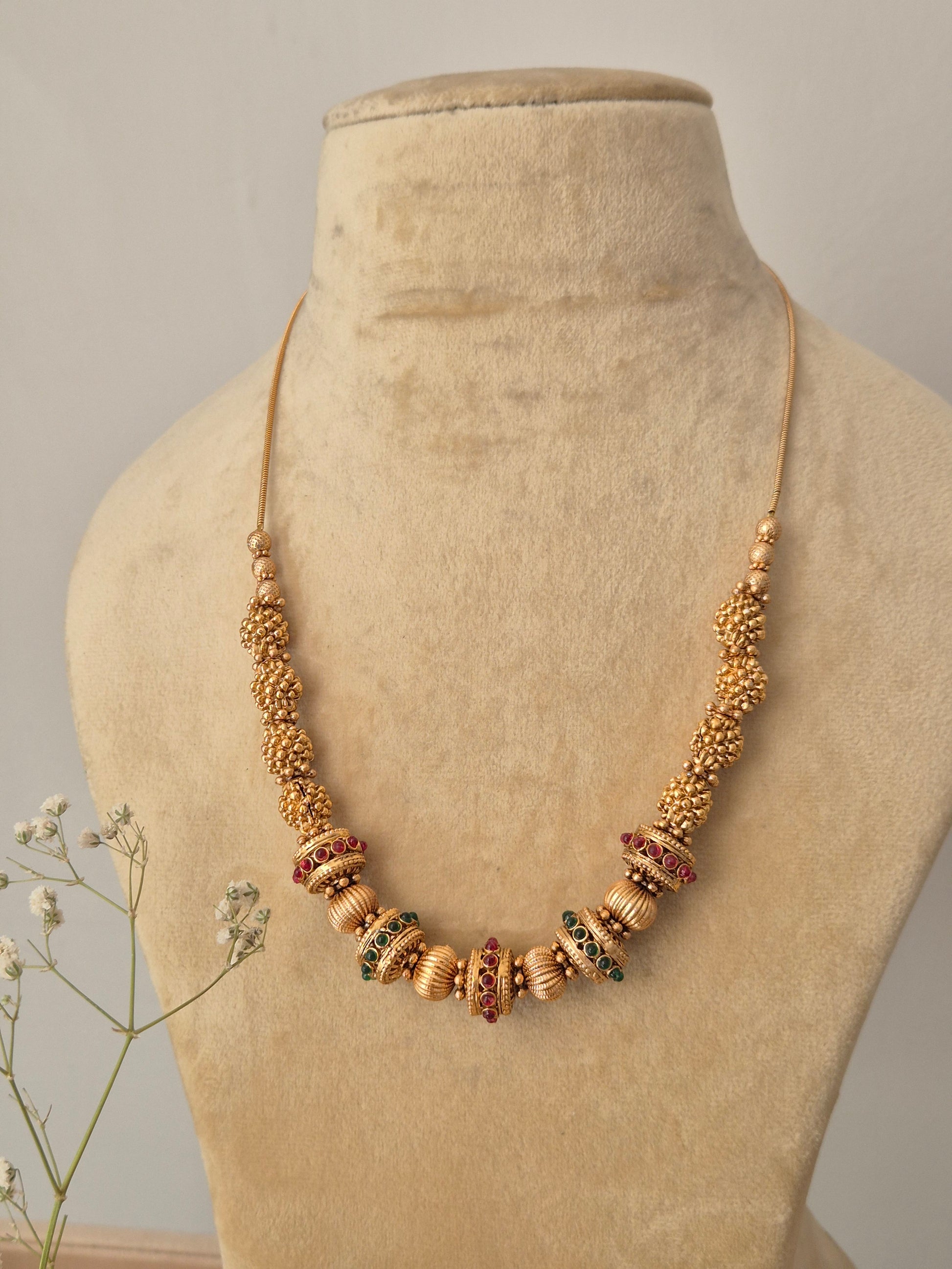 Antique gold-style Devi necklace with colorful stone embellishments, styled on a neutral bust with baby's breath flowers for a modern Indian aesthetic by naitri.in