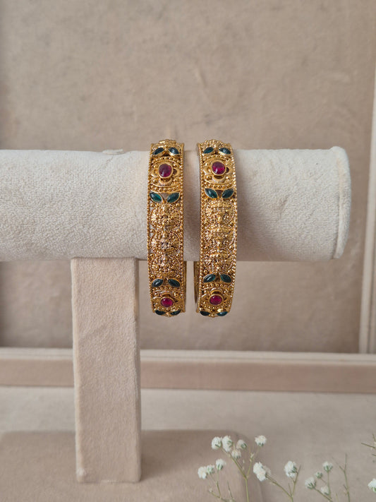 Set of 2 Dhanashree gold bangles by Naitri featuring intricate traditional engravings, accented with alternating ruby-red and emerald-green gemstones, by naitri.in