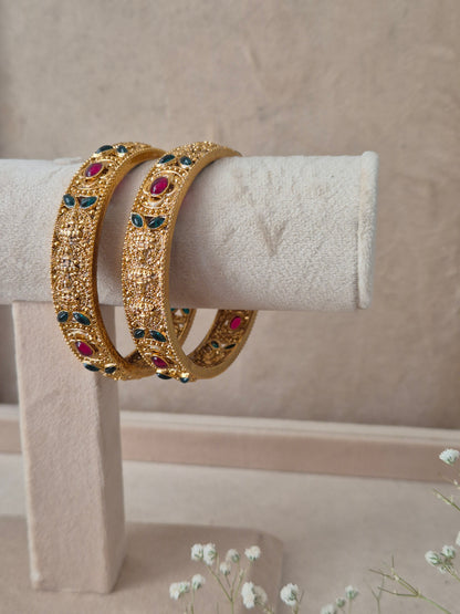 Pair of Naitri's Dhanashree gold bangles showcasing detailed ethnic craftsmanship, embedded with vibrant pinkish-red and green accent stones, by naitri.in