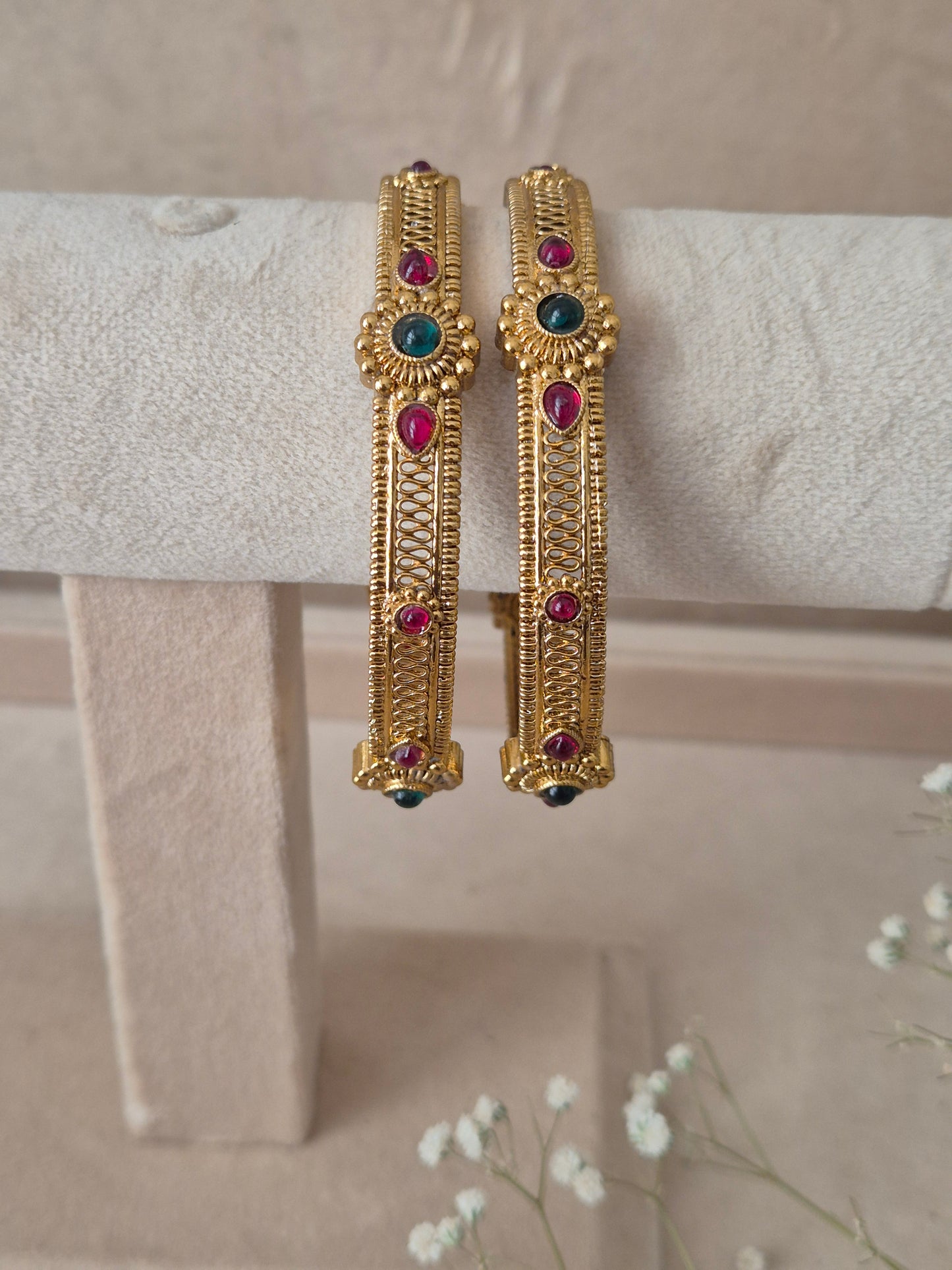 Pair of Naitri's Dhruvi gold bangles showcasing detailed ethnic craftsmanship, embedded with vibrant pinkish-red and green accent stones, by naitri.in