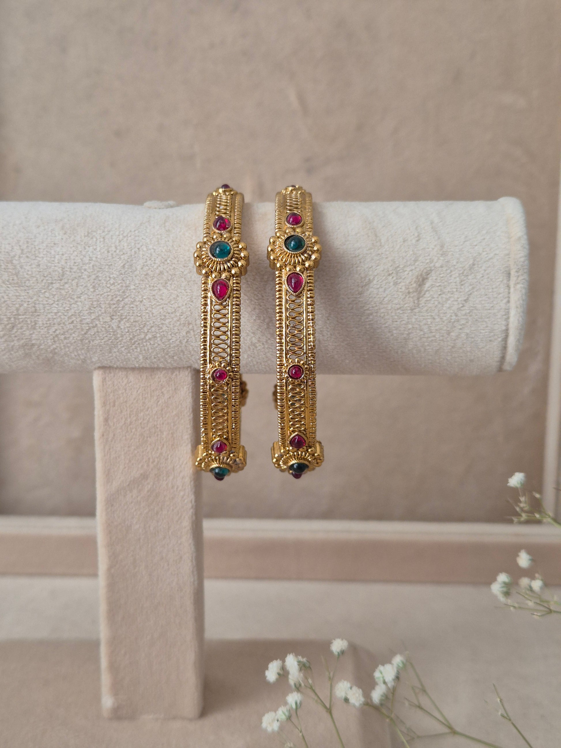 Set of 2 Dhruvi gold bangles by Naitri featuring intricate traditional filigree carvings, accented with alternating ruby-red and emerald-green gemstones by naitri.in