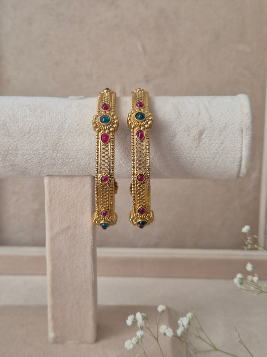 Set of 2 Dhruvi gold bangles by Naitri featuring intricate traditional filigree carvings, accented with alternating ruby-red and emerald-green gemstones by naitri.in