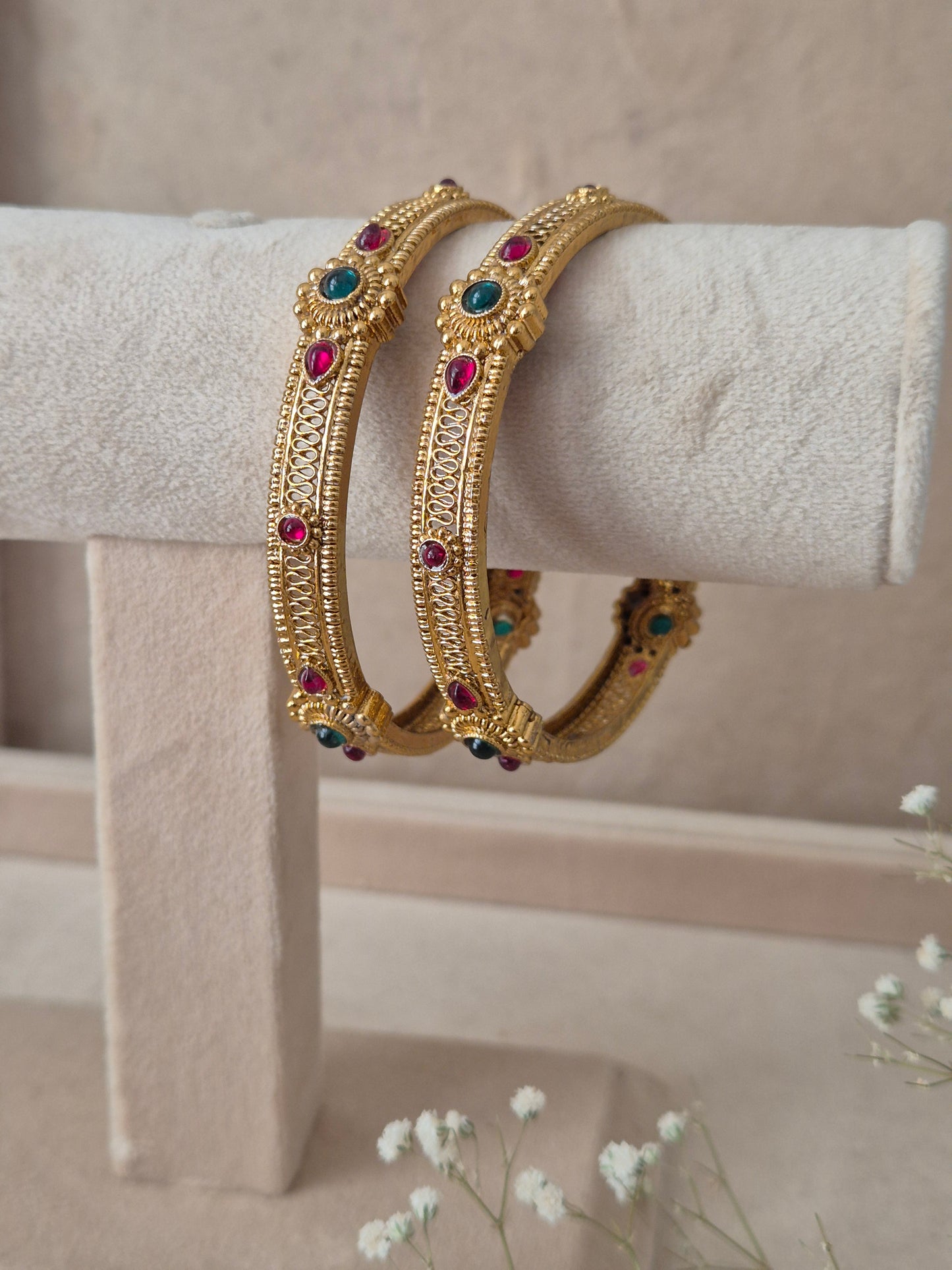 Two traditional gold-plated ethnic bangles with detailed textured patterns and inlaid red and green gemstones by naitri.in