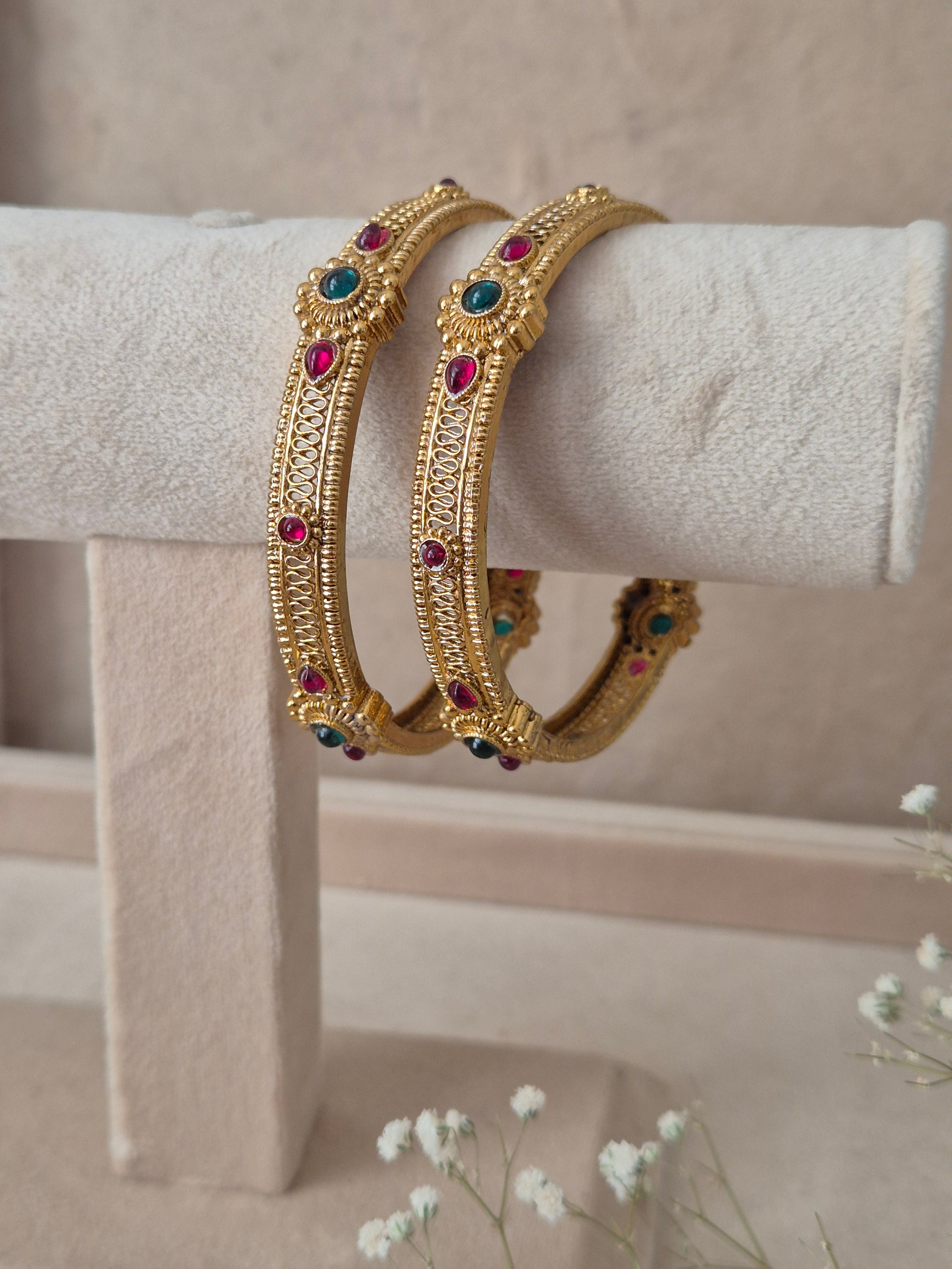 Two traditional gold-plated ethnic bangles with detailed textured patterns and inlaid red and green gemstones by naitri.in