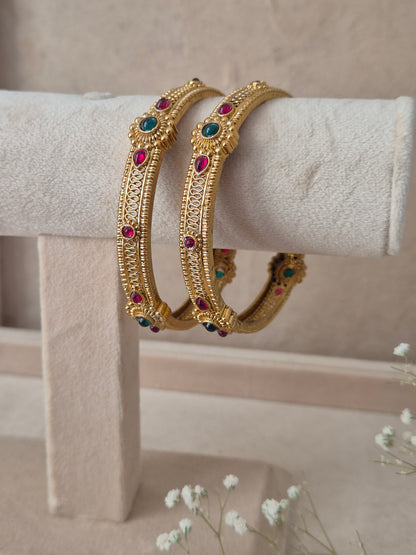 Two traditional gold-plated ethnic bangles with detailed textured patterns and inlaid red and green gemstones by naitri.in