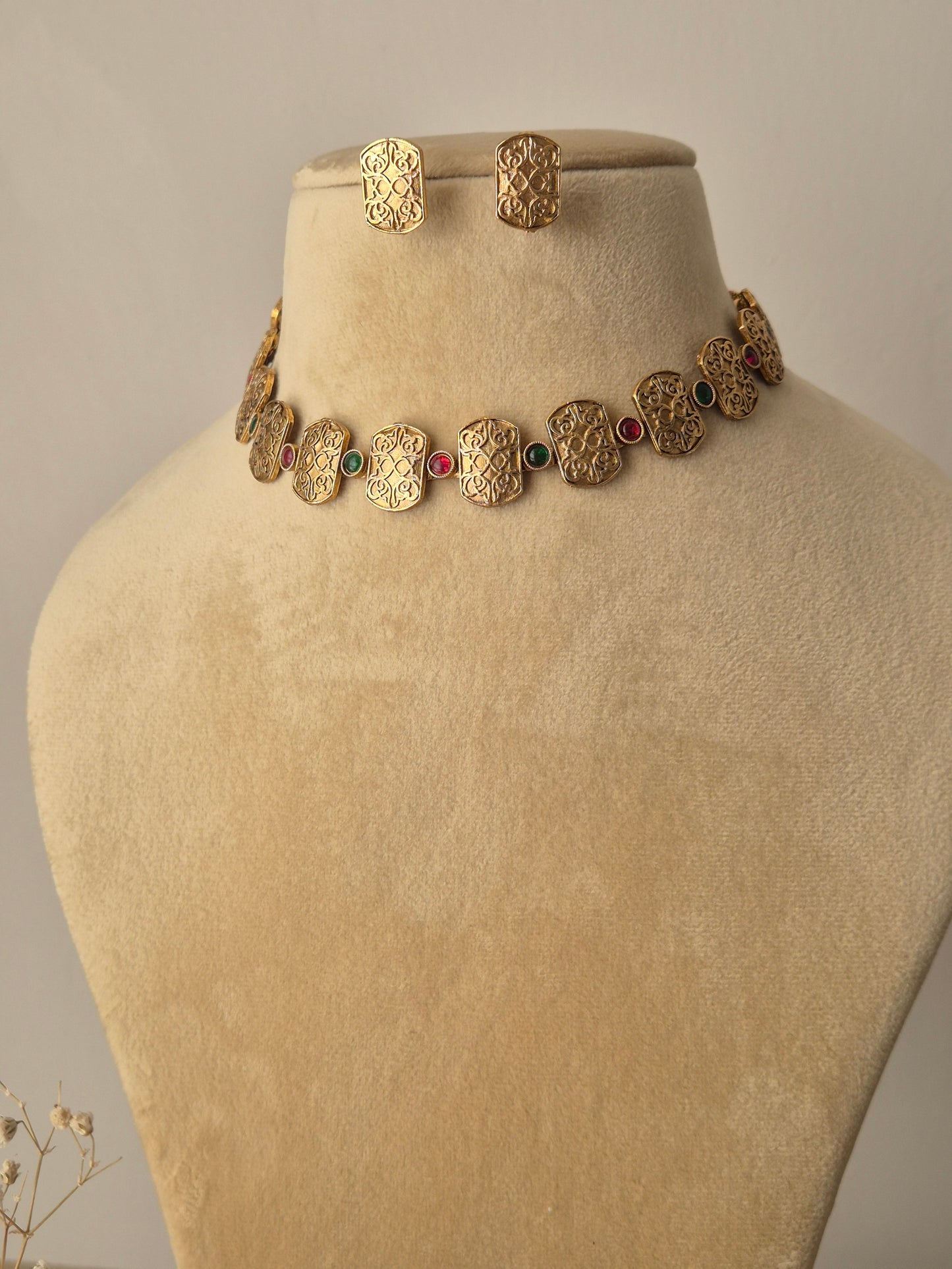 Close-up of the Diksha Necklace Set: a gold-toned choker with alternating engraved plates and small red and green stones, paired with rectangular matching studs by naitri.in