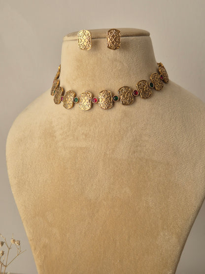 Close-up of the Diksha Necklace Set: a gold-toned choker with alternating engraved plates and small red and green stones, paired with rectangular matching studs by naitri.in