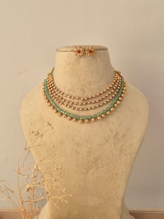 Divyanshi Multi-Strand Choker Set displayed on a beige mannequin, featuring layered metallic gold beads combined with elegant green crystal strands and matching floral stud earrings by Naitri.in