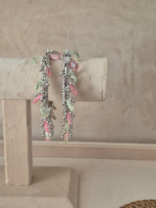 Set of two oxidized silver Eartha bangles hanging from a beige velvet display stand, featuring a row of dangling, faceted pastel pink and mint green teardrop gemstones and small metallic beads by naitri.in