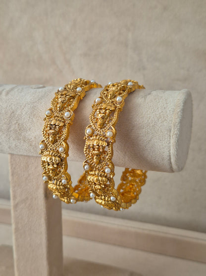 Elegant golden statement bangles with pearl embellishments exhibited on a neutral luxury jewelry display next to small white flowers by naitri.in