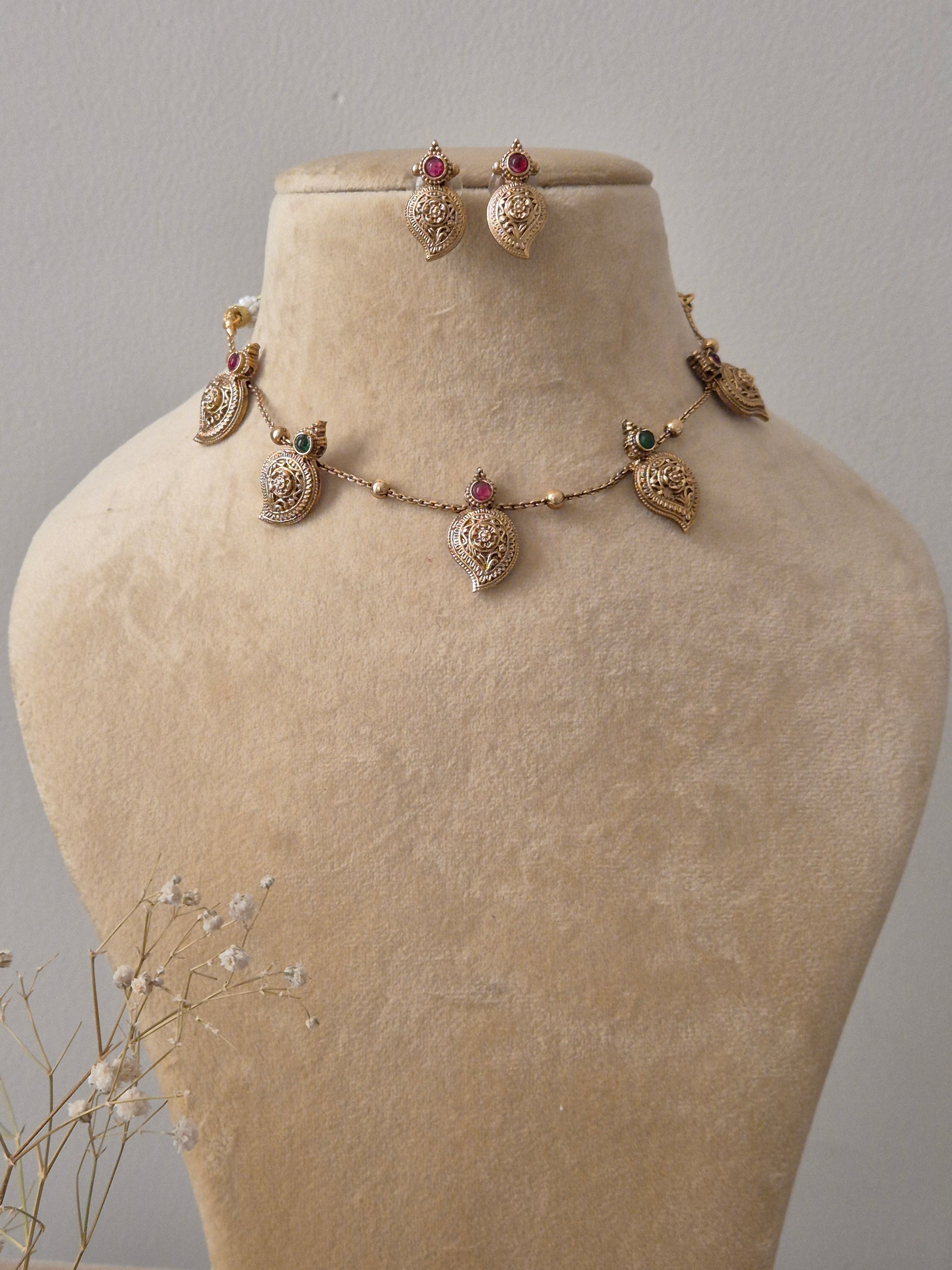 Set by Naitri featuring a gold-plated brass chain with five leaf-shaped pendants, red and green stone accents, and matching leaf-shaped stud earrings