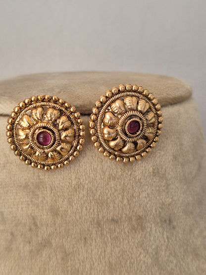 A pair of ornate gold-toned circular earrings with intricate floral detailing and a small dark red center stone, by naitri.in