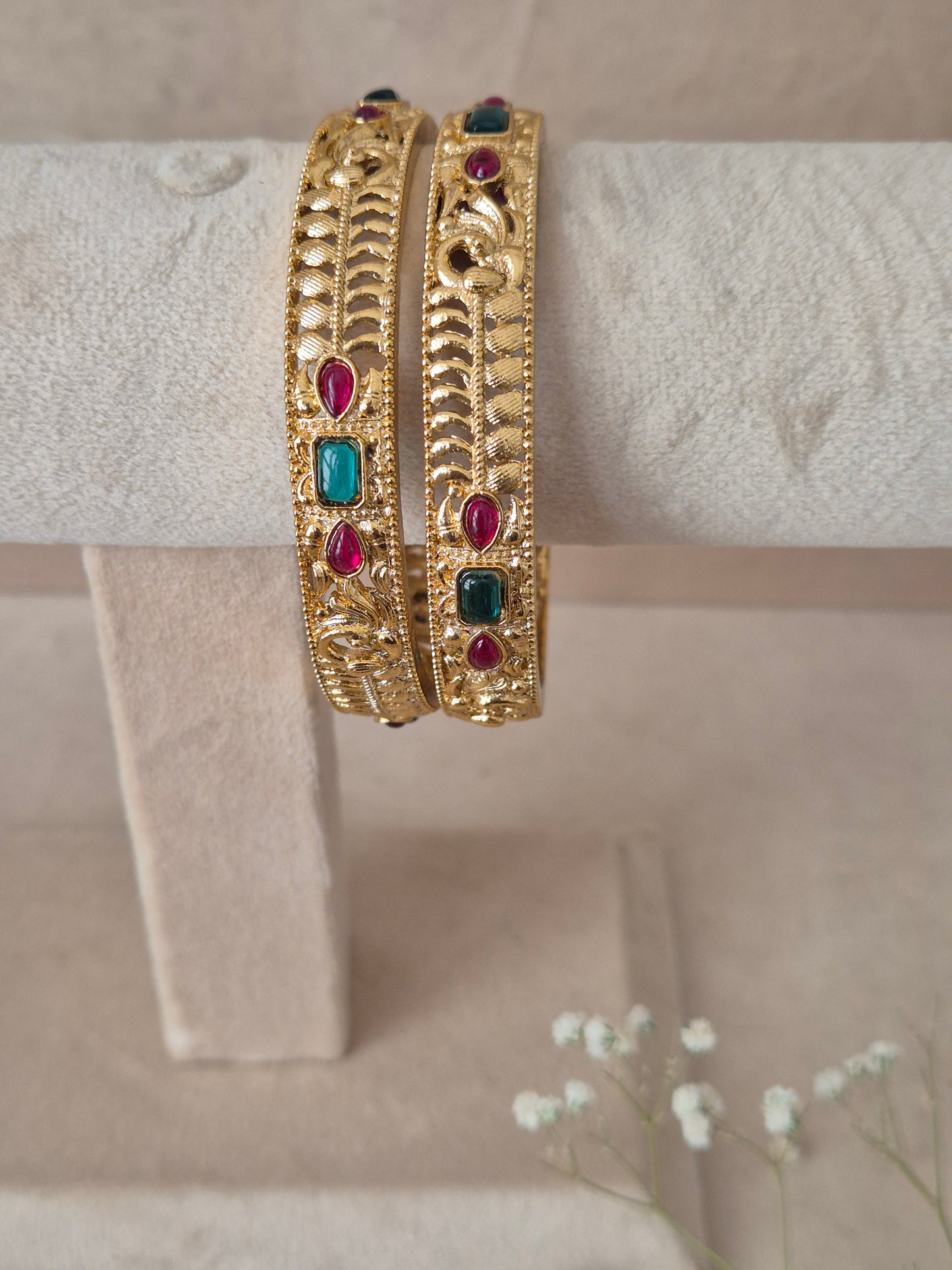 Set of 2 Esha gold bangles by Naitri featuring intricate traditional textured engravings, accented with alternating ruby-red and emerald-green gemstones by naitri.in