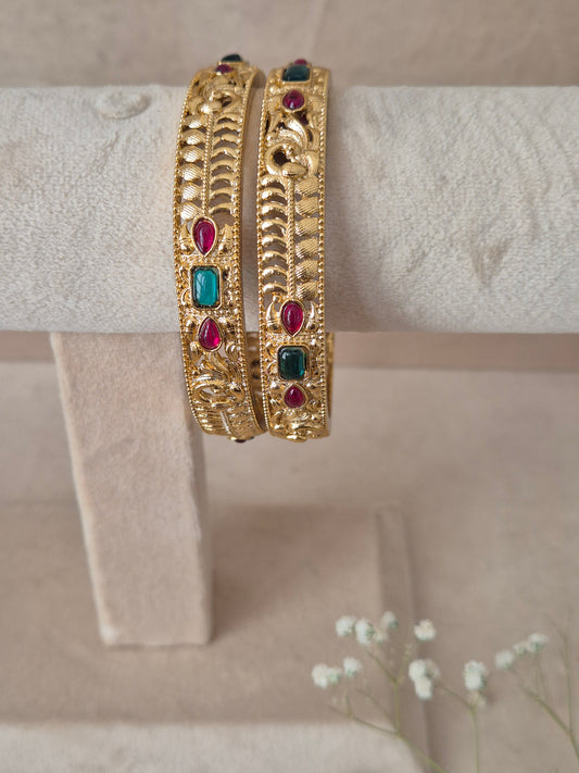 Set of 2 Esha gold bangles by Naitri featuring intricate traditional textured engravings, accented with alternating ruby-red and emerald-green gemstones by naitri.in