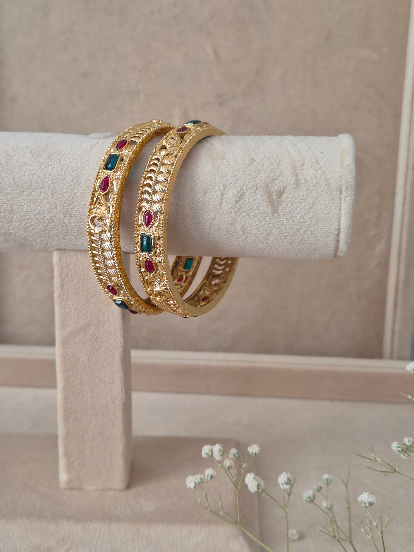Two traditional gold-plated ethnic bangles with detailed geometric patterns and inlaid red and green gemstones by naitri.in