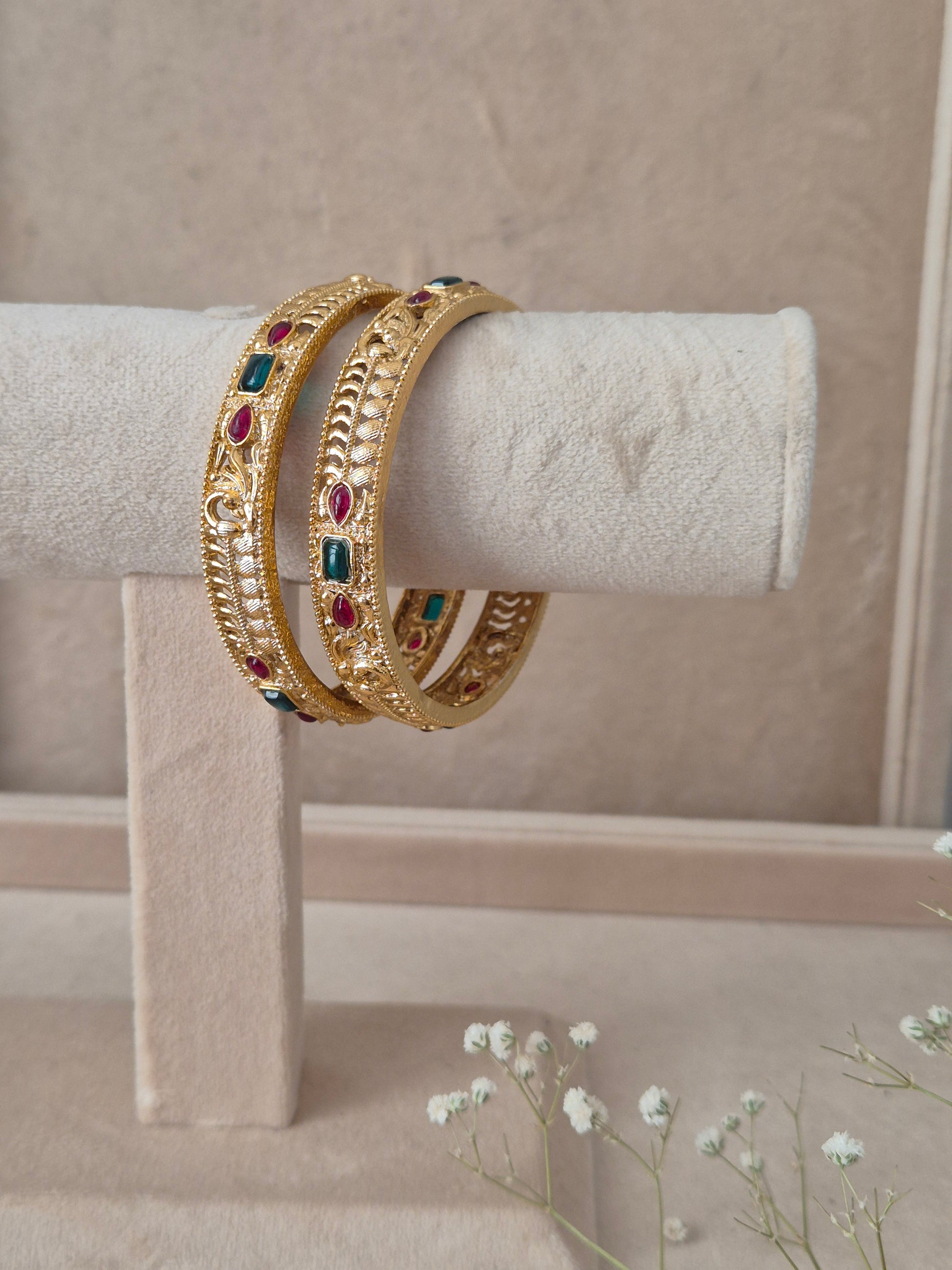 Two traditional gold-plated ethnic bangles with detailed geometric patterns and inlaid red and green gemstones by naitri.in