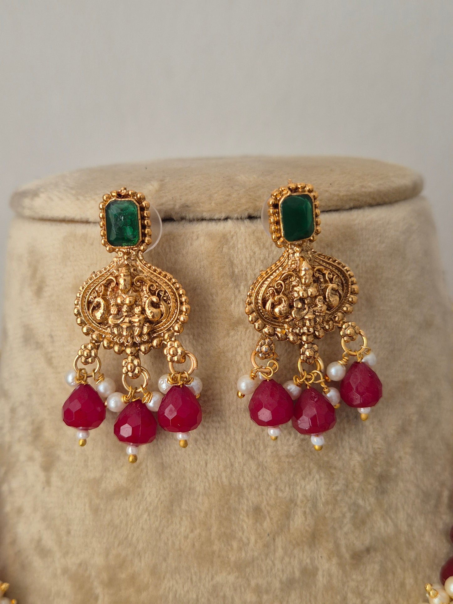 A pair of ornate gold-toned traditional earrings with rectangular green stones, small white pearl-like beads, and three faceted red heart-shaped drops, by naitri.in