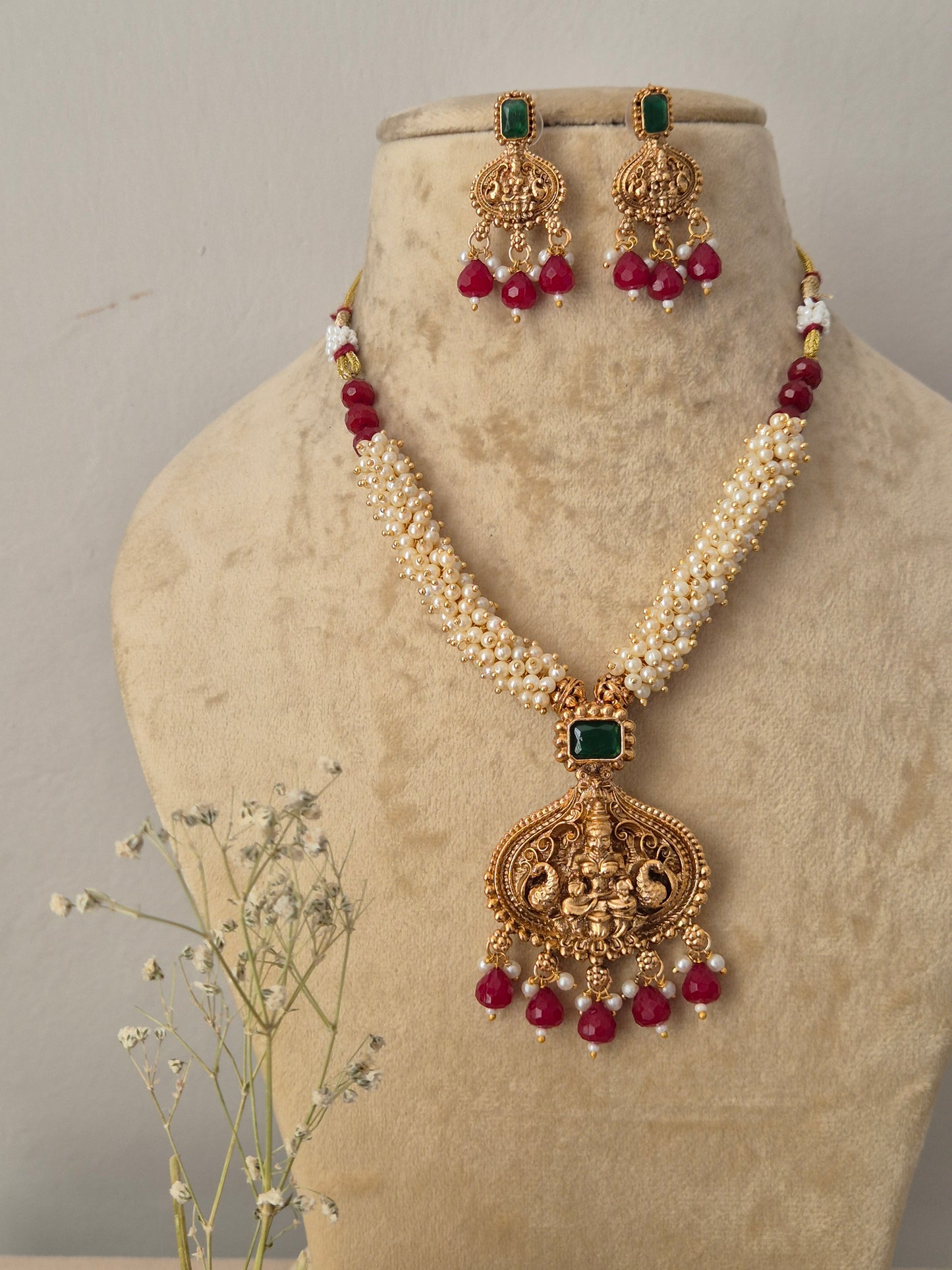 Gold-toned traditional Indian jewelry set featuring a multi-strand white pearl chain necklace with an intricate temple-style pendant, the pendant and matching hanging earrings are decorated with a square green gemstone and finished with a fringe of dangling red beads, by naitri.in