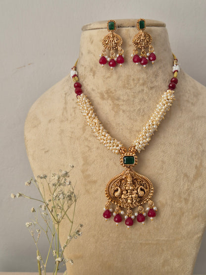 Gold-toned traditional Indian jewelry set featuring a multi-strand white pearl chain necklace with an intricate temple-style pendant, the pendant and matching hanging earrings are decorated with a square green gemstone and finished with a fringe of dangling red beads, by naitri.in