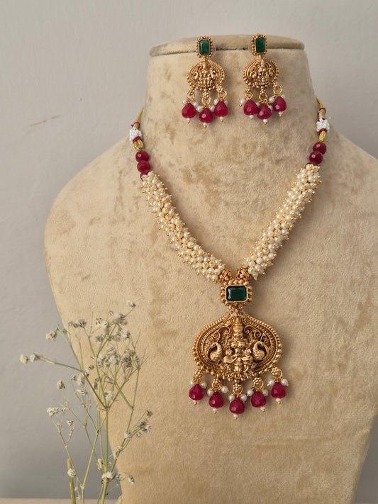 Gold-toned traditional Indian jewelry set featuring a multi-strand white pearl chain necklace with an intricate temple-style pendant, the pendant and matching hanging earrings are decorated with a square green gemstone and finished with a fringe of dangling red beads, by naitri.in