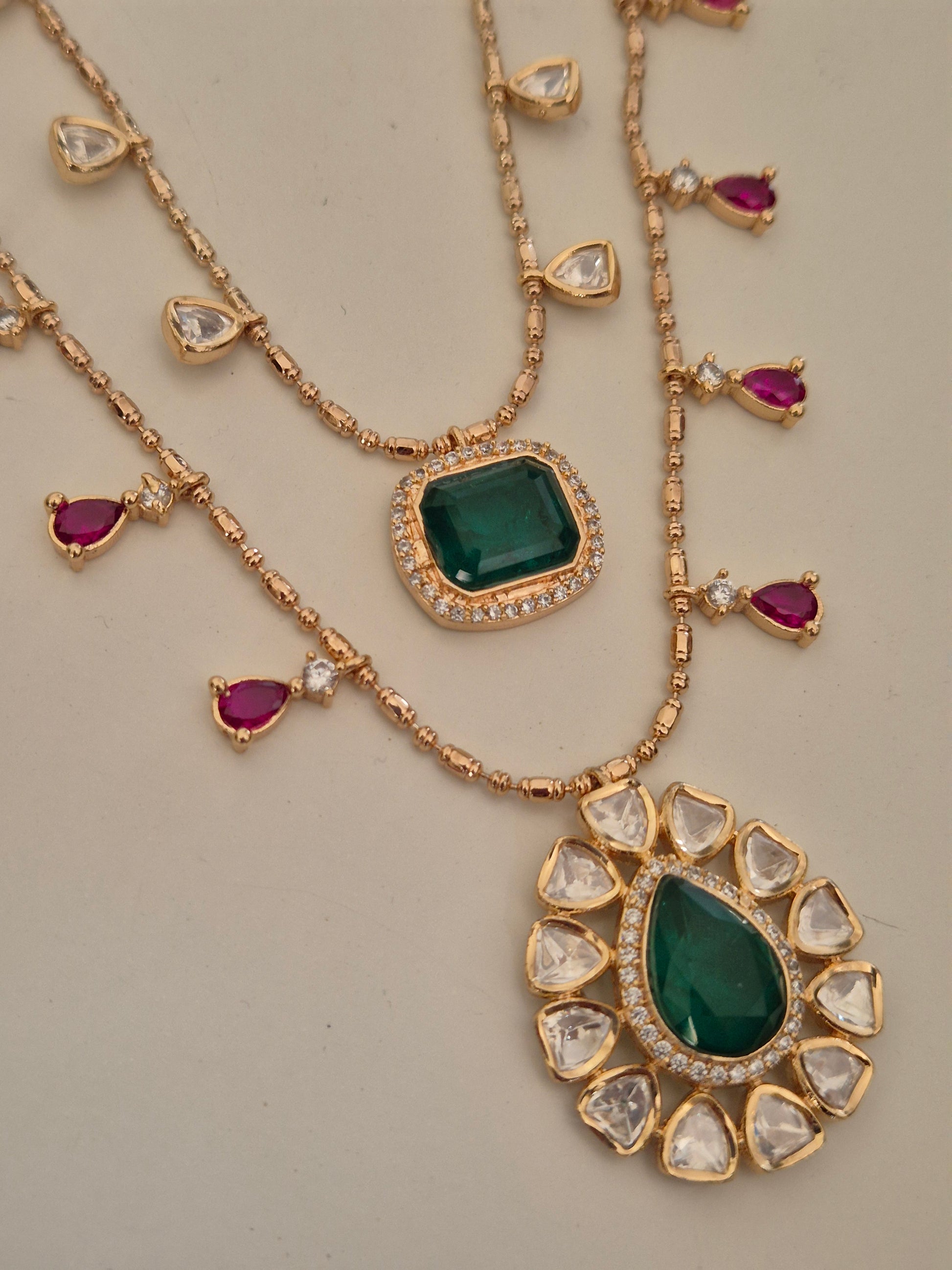 Rose-Gold polished necklace set featuring two tiers of American Diamond chains with central green emerald-style pendants and dangling ruby-pink and clear stones by naitri.in
