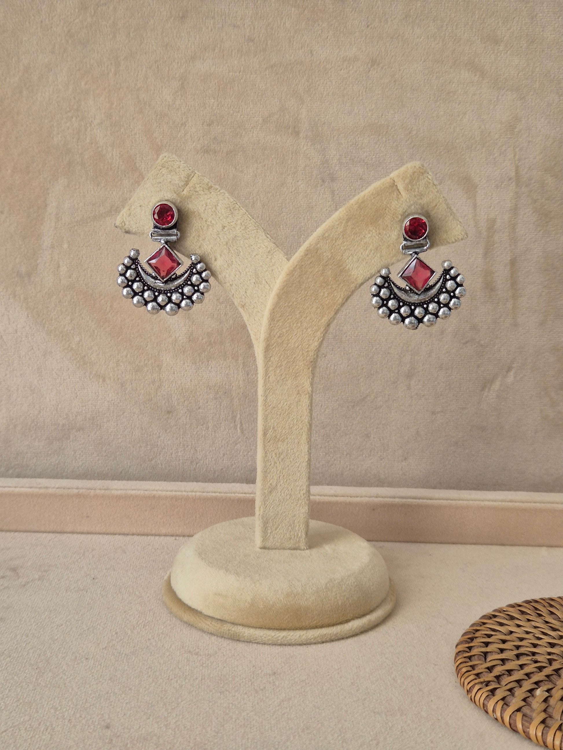 Pair of silver earrings with red stones on a beige stand Pair of Falguni silver oxidized drop earrings with a central red stone and a curved silver bead accent by naitri.in