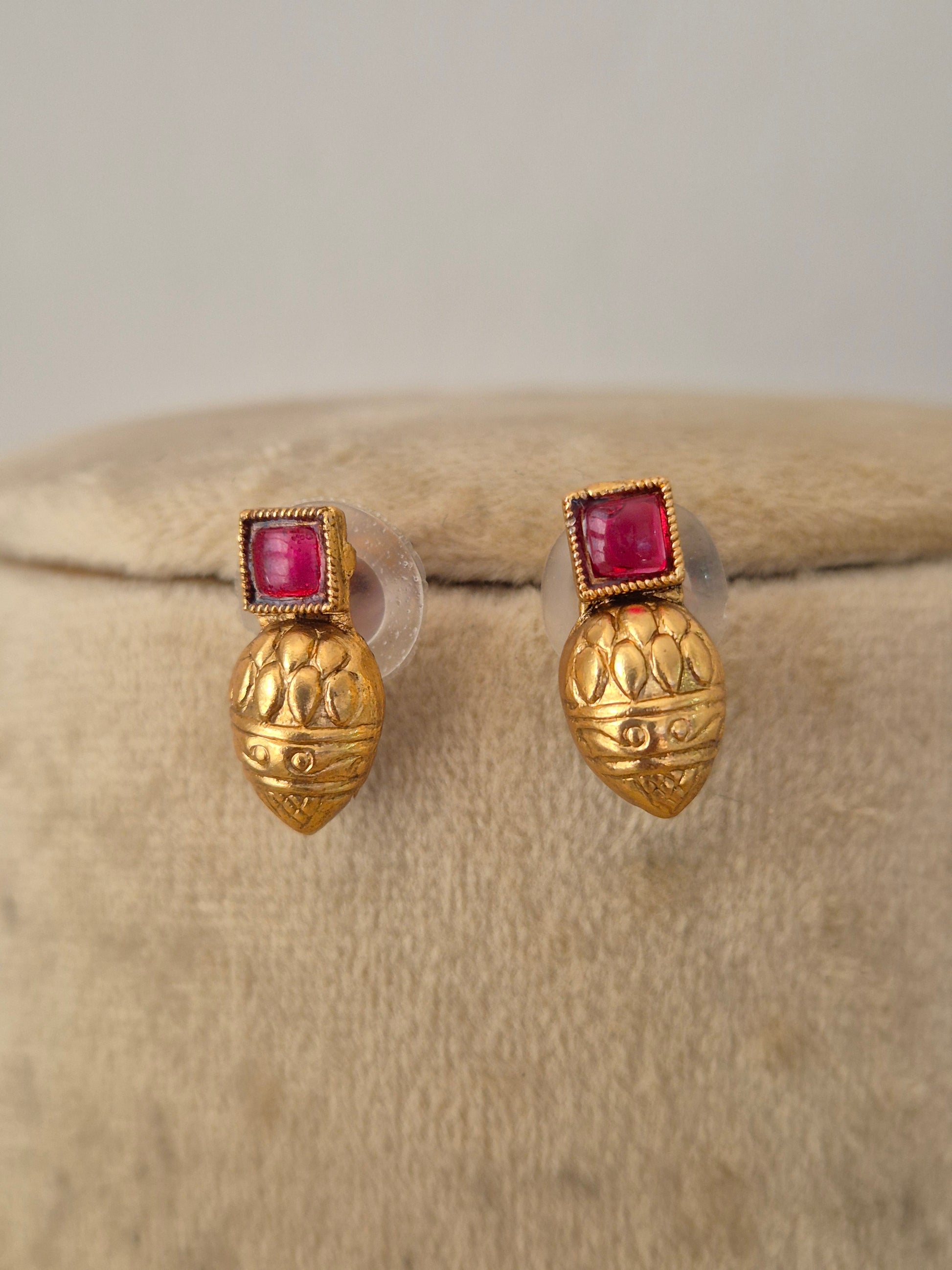 A pair of gold post earrings with a textured, lotus-like gold top and a square pinkish-red gemstone in a beaded border setting, by naitri.in