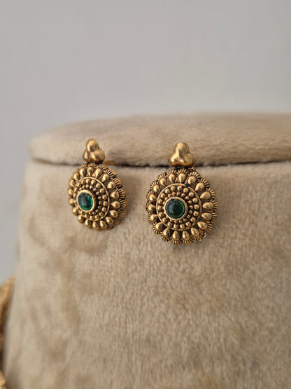 Antique gold-toned traditional circular earrings feature intricate, textured sunflower-like metallic patterns surrounding a central deep green gemstone by naitri.in
