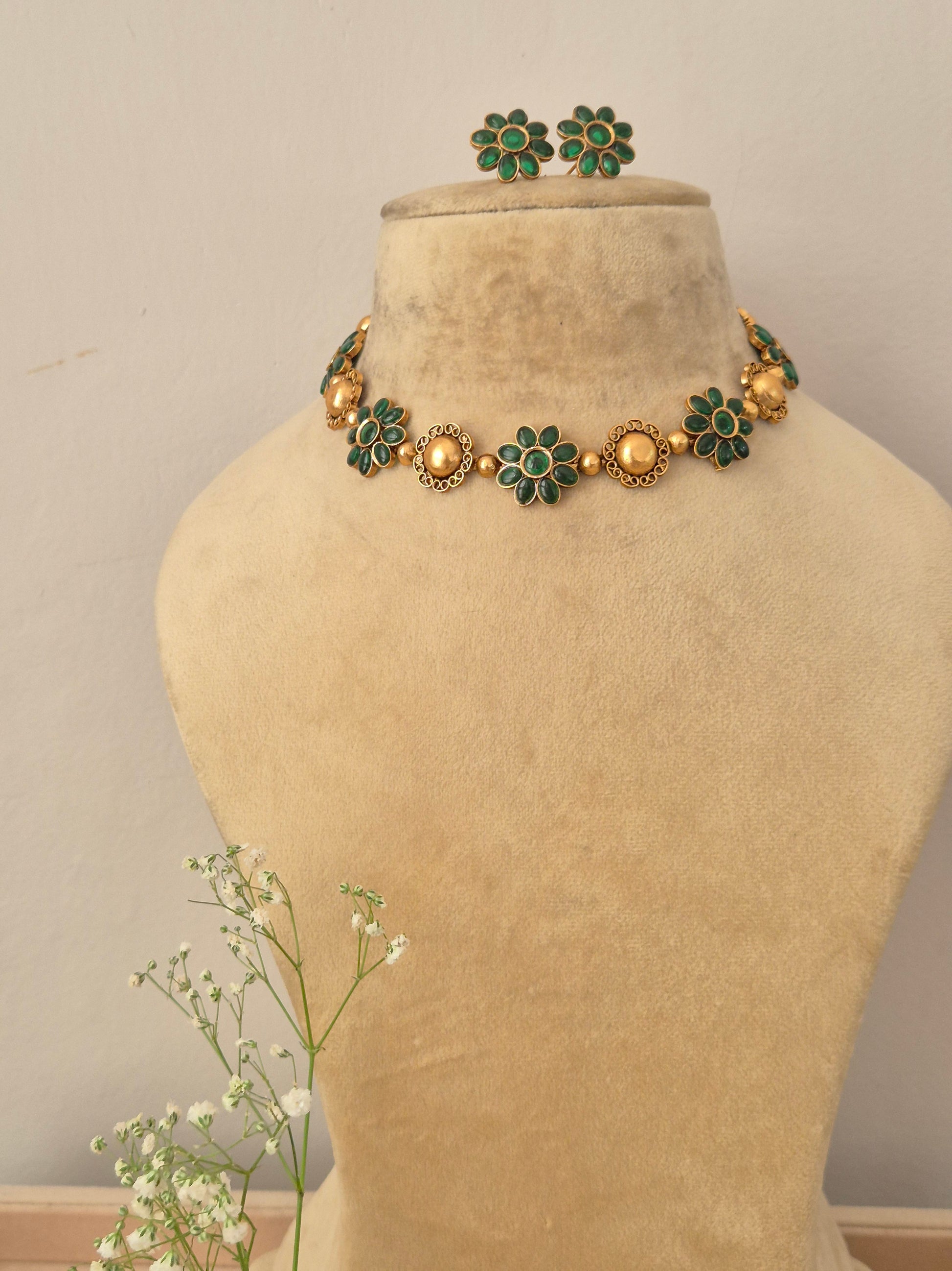 A gold-plated ethnic choker necklace featuring alternating textured round gold charms and green gemstone floral motifs,with matching flower stud earrings by naitri.in