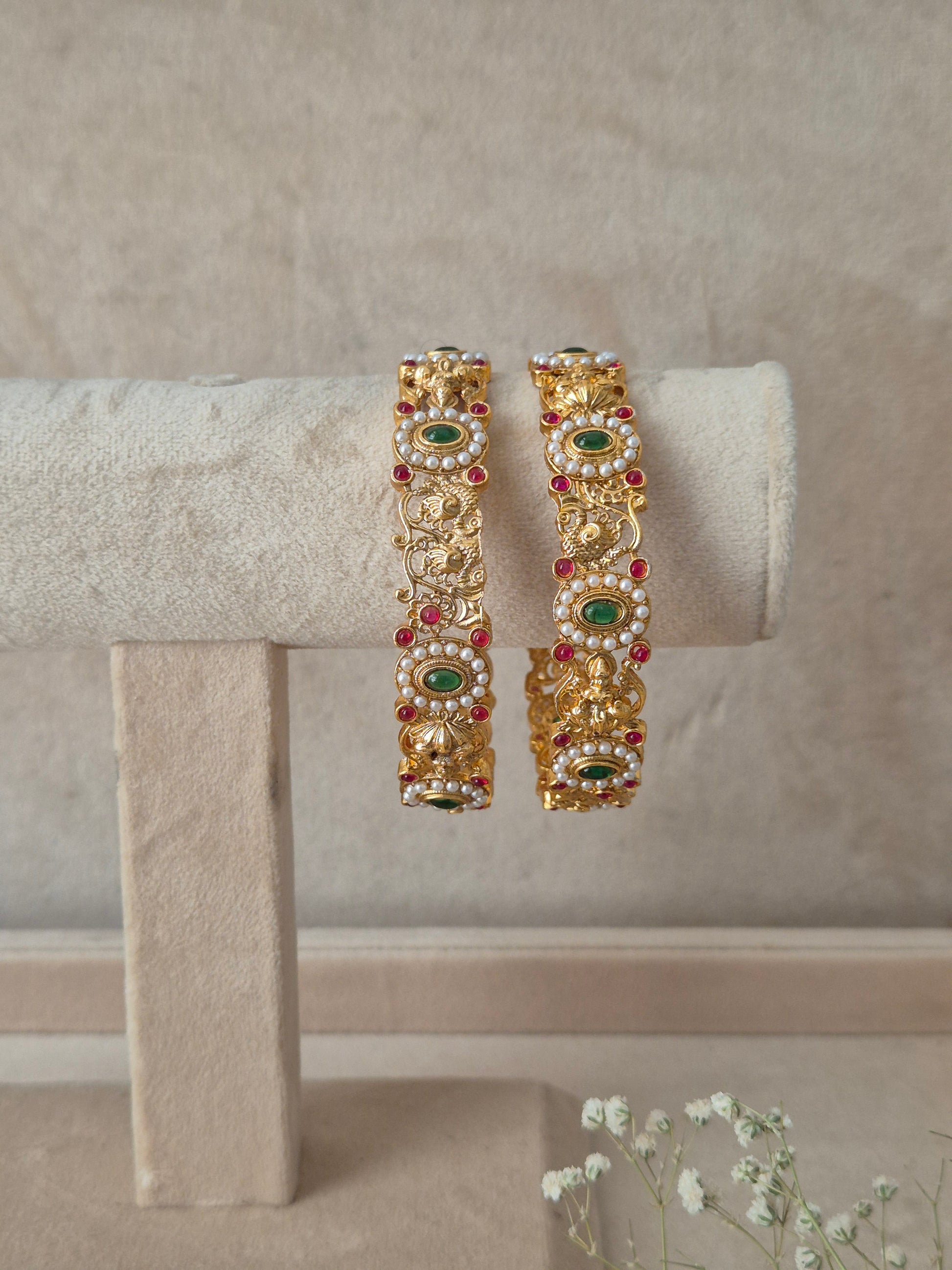 A set of two gold-plated Gaytri bangles featuring intricate floral and vine metalwork inlaid with alternating red and green gemstone by naitri.ihn