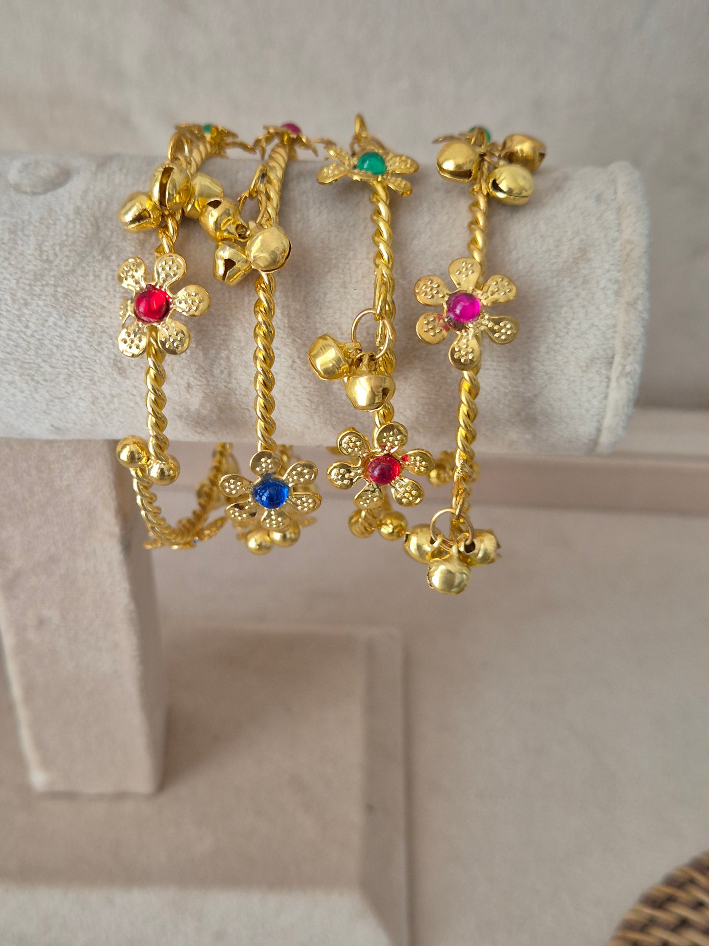 Set of four gold-plated Geetanjali bangles hanging on a beige velvet display stand, featuring delicate floral charms inlaid with ruby red, emerald green, and sapphire blue stones, finished with dangling gold chains and tiny ghungroo beads by naitri.in