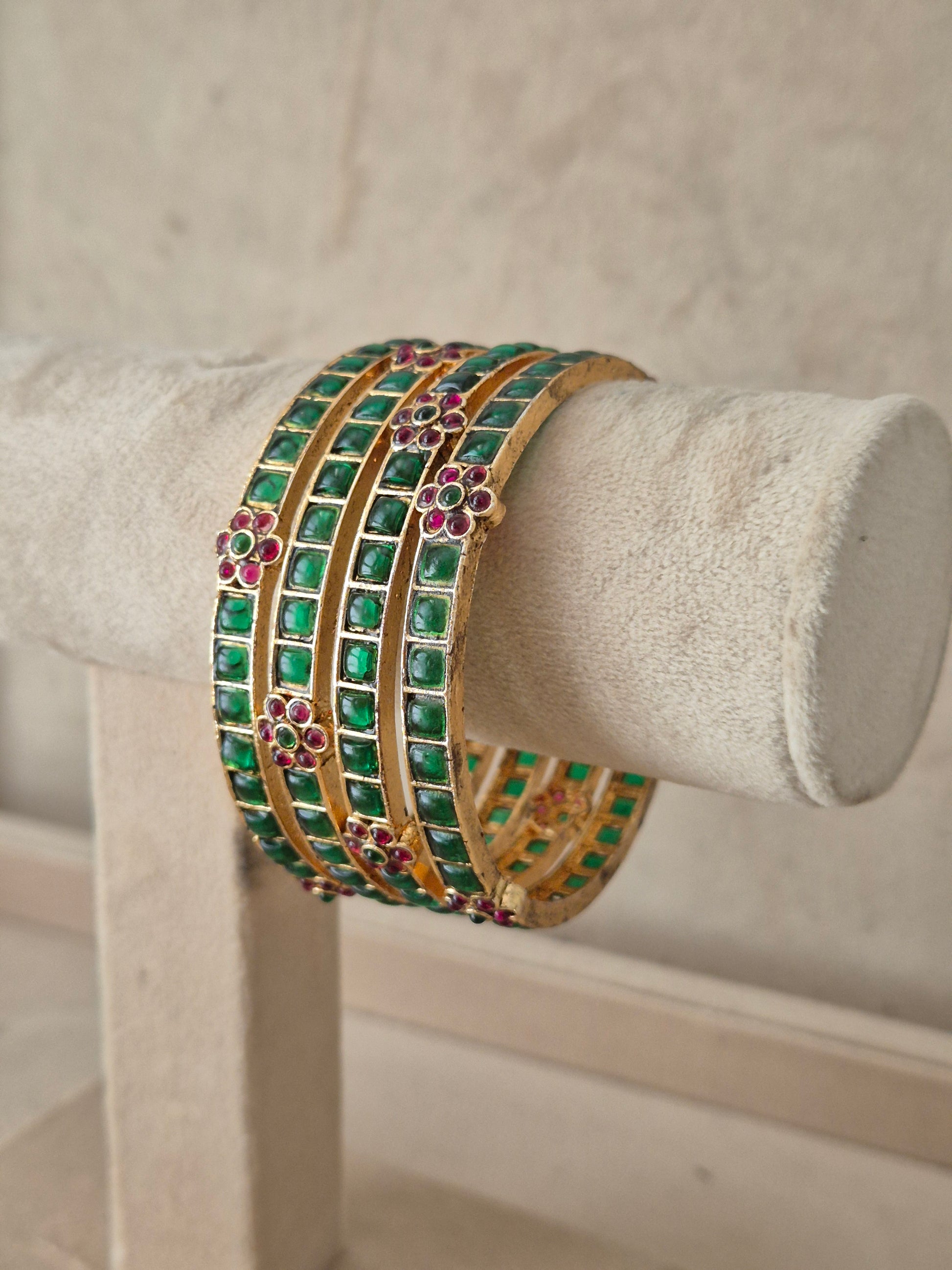 A stack of four gold bangles adorned with green square stones and pink floral accents, Traditional Indian festive bangles adorned with green gemstones and pink floral stone clusters by naitri.in