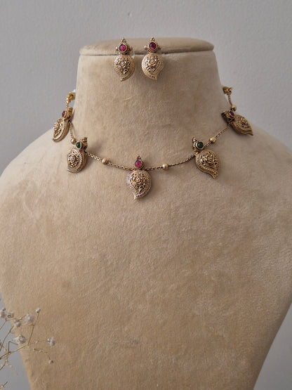 Gold-toned ethnic jewelry set with intricate leaf-motif charms, ruby-red and emerald-green stones, and tiny gold bead detailing by naitri.in