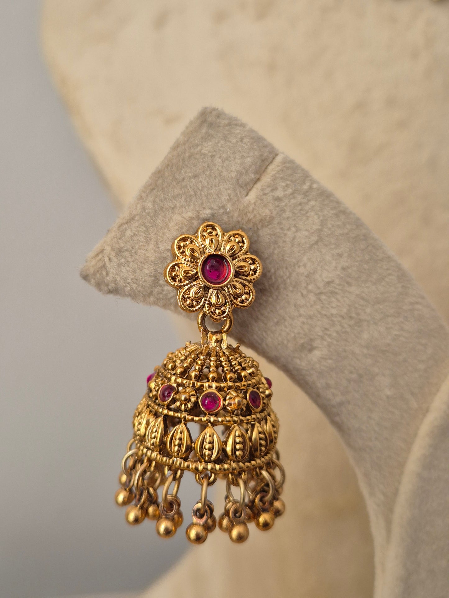 An ornate gold-toned Indian jhumka earring with a central pink gemstone and small pink accent stones, the matching bell-shaped jhumka earrings are detailed with floral studs and hanging gold bead clusters by naitri.in