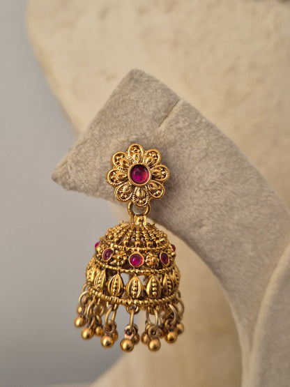 An ornate gold-toned Indian jhumka earring with a central pink gemstone and small pink accent stones, the matching bell-shaped jhumka earrings are detailed with floral studs and hanging gold bead clusters by naitri.in