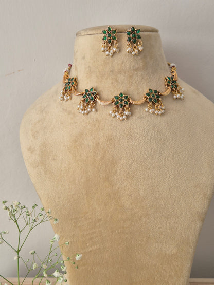 A gold-plated choker necklace featuring repeating flower motifs with emerald-green stones and hanging pearl clusters with matching earrings by naitri.in